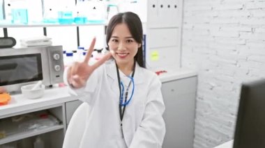 Laboratuvardaki kendine güvenen genç Çinli bilim kadını zaferle ışıldıyor, iki parmağıyla bariz bir 
