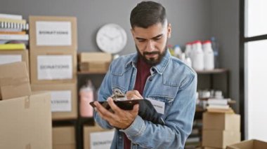 Kot ceket giyen sakallı bir genç gönüllü olarak bağış kutularıyla dolu bir depoda not defterine yazıyor..