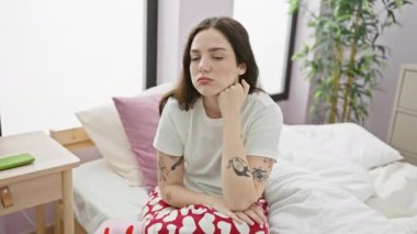 Pijamalı genç bir kadın yatakta yorgun ve sıkkın bir şekilde oturuyor, depresyonda, yatak odasında düşüncelerini değiştiriyor.