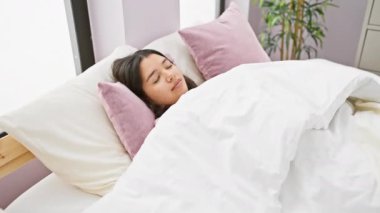 Genç bir İspanyol kadın gülümseyerek yatakta uzanıyor, güzel ışıklandırılmış bir yatak odasında rahat bir sabah rutinini canlandırıyor..
