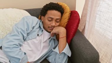 Kıvırcık saçlı yakışıklı bir genç, günlük kıyafetler giyiyor, yorgun bir şekilde kanepede oturuyor ve içeride esniyor..