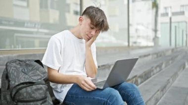 Stresli, sarı saçlı beyaz bir erkek öğrenci şehir kampüsünde bir bankta dizüstü bilgisayarını kullanarak oturuyor ve hayal kırıklığını gösteriyor..