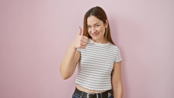 Young hispanic woman with blue eyes standing smiling happy and positive, thumb up doing excellent and approval sign over isolated pink background