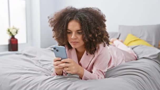 カーリーヘアの陽気なヒスパニック女性はベッドに横たわり スマートフォンでテキストをしながら承認の親指サインを与えます — ストック動画