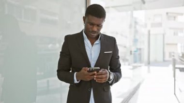Kendine güvenen Afrikalı takım elbiseli adam güneşli şehir caddesinde akıllı telefon kullanıyor.