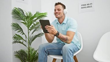 Beyaz modern bekleme odasında bitki ve ücretsiz kablosuz ağ tabelalı tablet kullanan, kulaklıklı gülen İspanyol adam..