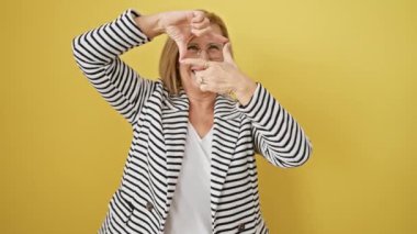 Sarışın, orta yaşlı kadın çizgili ceketli, elleri çerçeveli sarı bir duvara yaslanmış, parmakları fotoğraf oluşturuyor. Mutlu yüzü yaratıcılık ve fotoğrafçılık kavramını somutlaştırıyor..