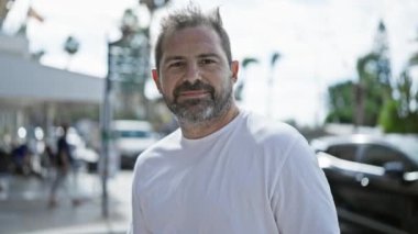 Gülümseyen orta yaşlı, gri saçlı İspanyol bir adam güneşli bir şehir sokağında baş parmağını sallıyor..