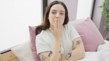 Pijamalı sevimli genç bir kadın kameraya doğru seksi bir öpücük yolluyor. Kendinden emin görünüyor, yatak odasında oturuyor, havada basit bir el hareketiyle ışıltılı aşkı ifade ediyor..