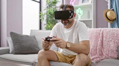 Modern bir oturma odasında video oyunları oynayan sakallı ve kulaklıklı olgun bir adam.