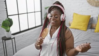 Neşeli Afrikalı Amerikalı kadın mikrofon gibi fırçayla şarkı söyleyerek eğleniyor, yatağında oturuyor, rahat yatak odasında müzik dinliyor.