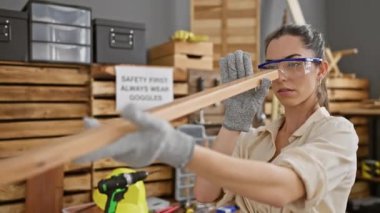Güzel, genç İspanyol kadın marangoz marangoz, marangozluk atölyesinde tahtayı ustalıkla inceliyor, rahat bir tavırla koruyucu gözlük takıyor.