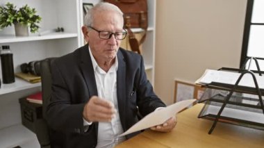 Seçkin kıdemli adam işine odaklanmış, iş belgelerini okuyor, ofiste rahat bir şekilde gözlüklerini çıkarıyor.