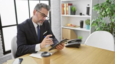 Çekici, gri saçlı, genç İspanyol bir adam, ofisinde tamamen işine odaklanmış, dokunmatik deftere notlar alıyor, sakallı, gözlüklü ve takım elbiseli yakışıklı bir profesyonel, kapalı bir masada oturuyor.