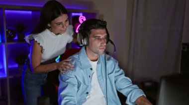 Bir kadın, akşamları neon ışıklı oyun odasında oturan bir adamın yanında duruyor. Ev ortamında eğlence ve teknolojinin göstergesi olarak..