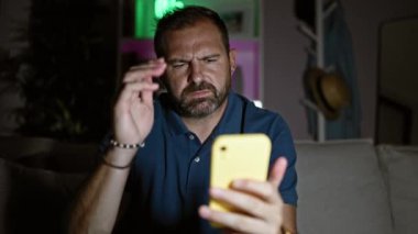 Baş ağrısı olan olgun bir adam gece geç saatlerde evde akıllı telefon kullanıyor.