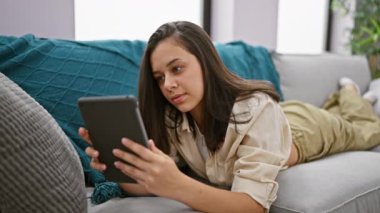 Evdeki kanepede uzanmış dokunmatik ped kullanan genç, güzel İspanyol kadın.