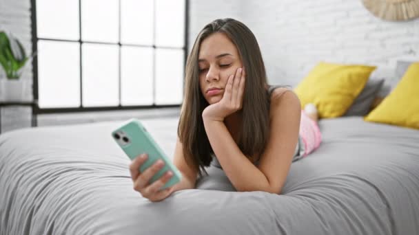 疲れ果てた若いヒスパニック系女性がベッドに横たわり 居心地の良い寝室でスマートフォンを使用しながらヨーイング — ストック動画