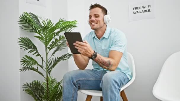 Usmívající Hispánský Muž Sluchátky Pomocí Tabletu Bílé Moderní Čekárně Květinovým — Stock video