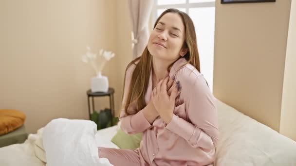 一个快乐的年轻女人 蓝眼睛 穿着睡衣坐在床上 带着感激的微笑 在她的房间里做着一个健康的早晨的姿势 双手放在胸前 — 图库视频影像