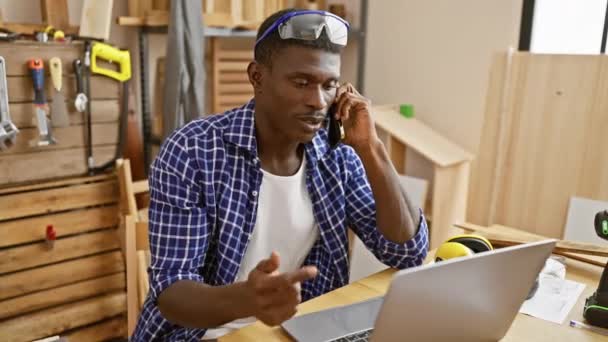ワークショップのアフリカ系アメリカ人男性 大工の道具に囲まれたノートパソコンで電話で — ストック動画