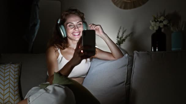 若い女性は自宅で夜を楽しんでおり スマートフォンやヘッドフォンを使用して友人とビデオ通話しています — ストック動画