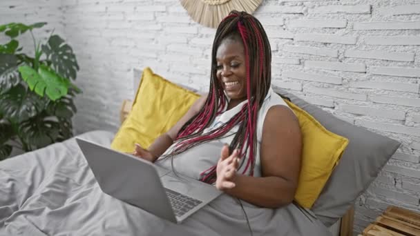 アフリカ系アメリカ人女性がベッドルームのベッドに座ってラップトップでビデオ通話をしている — ストック動画