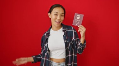 Neşeli genç Çinli kadın parmaklarıyla işaret ediyor, Çin pasaportu tutuyor, izole edilmiş kırmızı arka planda harika bir neşe duygusunu simgeliyor.