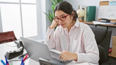 Gözlüklü, odaklanmış bir İspanyol kadın iyi organize edilmiş bir ofis ortamında dizüstü bilgisayarının başında çalışıyor..