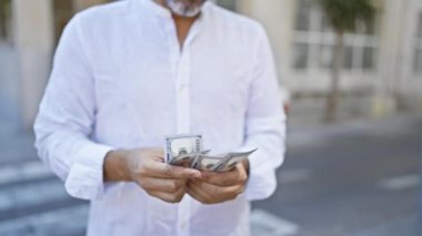 Genç, Latin asıllı, kır saçlı, gülümseyen, kendinden emin, sokakta dolar sayan bir adam.