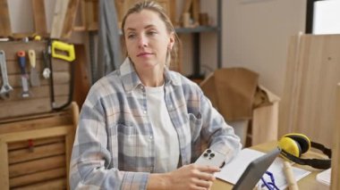 Marangozluk atölyesinde elinde aletlerle tablet kullanan gülümseyen beyaz bir kadın girişimci..