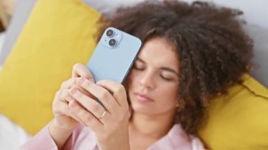 Evdeki yatak odasında akıllı telefon kullanan kıvırcık saçlı İspanyol kadın yaşam tarzı ve teknolojisini sergiliyor..
