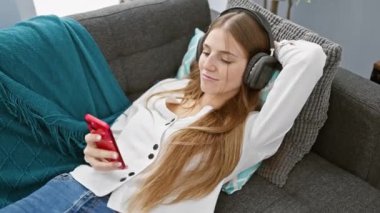 Genç bir kadın, kanepede, kulaklıkları ve akıllı telefonuyla rahat bir oturma odasında keyfine bakıyor..