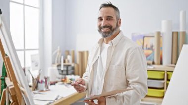 Parlak sanat stüdyosunda tuvale resim yapan gülümseyen sakallı adam.