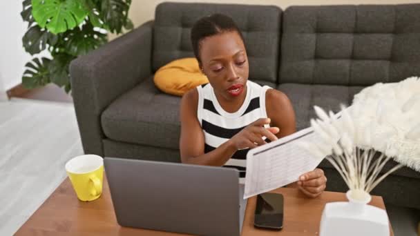 自宅で深刻なビデオ通話会議中に文書を読むことに集中したアフリカ系アメリカ人女性は リビングルームのソファーに快適に座り テクノロジーとの仕事のバランスを完璧に取っています — ストック動画