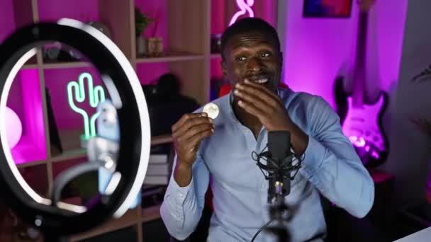 非洲男子在一个充满活力的播客演播室里展示比特币 晚上有霓虹灯和戒指灯 — 图库视频影像