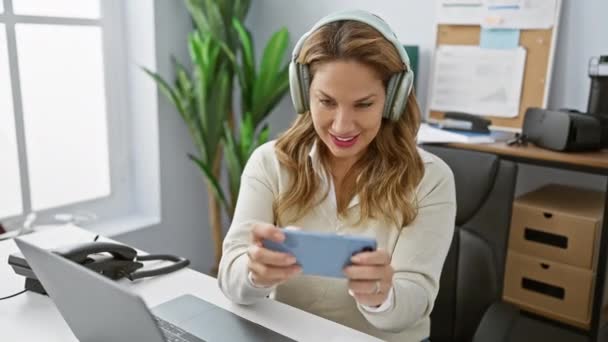 一个面带微笑的惊慌失措的女人喜欢现代办公室里带着耳机的手机游戏 — 图库视频影像