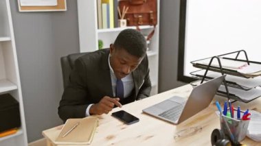 Ofis masasında dizüstü bilgisayarı, akıllı telefonu ve evrak işleriyle uğraşan sinirli bir Afrikalı.