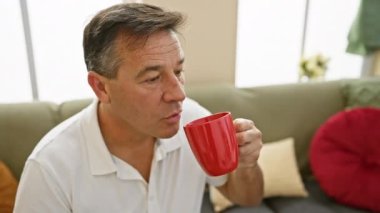Sıcak bir oturma odasında kırmızı bardaktan içen orta yaşlı bir adam günlük hayatı ve rahatlamayı simgeliyor..