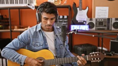 Bir kayıt stüdyosunda akustik gitar çalan yakışıklı, kulaklıklı ve mikrofonlu bir adam.
