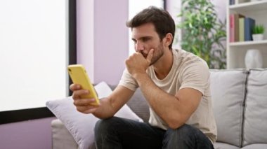 Akıllı telefon tutan düşünceli genç bir adam modern oturma odasında bir mesaj düşünüyor..