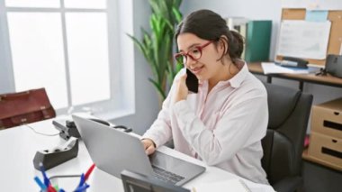 Genç bir İspanyol iş kadını telefonda gülümseyen ve dizüstü bilgisayar kullanan bir ofis ortamında birden fazla iş yapıyor..