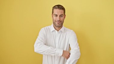 Neşeli genç İspanyol adam, kollarını kavuşturmuş, mutlu bir şekilde gülümsüyor, pozitiflik saçıyor. İzole edilmiş sarı bir duvara karşı doğal, dostane bir portre. Ne adam ama.!
