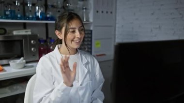 Beyaz laboratuvar önlüklü gülümseyen Latin kadın laboratuvar ortamında bilgisayar başında çalışıyor.