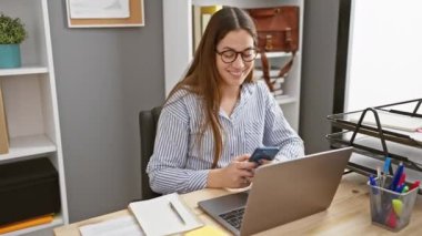 Ofiste dizüstü bilgisayarı, masası, gözlüğü ve sabit telefonu olan gülümseyen bir kadın.