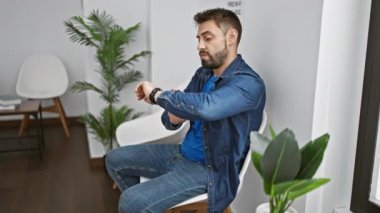 Stresli genç İspanyol adam, umutsuzca saati kontrol ediyor, bekleme odasında endişeyle oturuyor, baskı sorununu somutlaştırıyor.