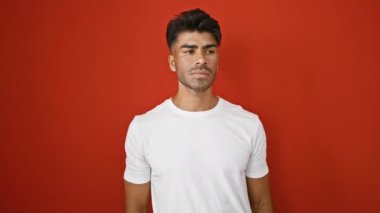 Sakallı yakışıklı genç adam gülümsüyor ve kırmızı duvarın arkasında el sallıyor..