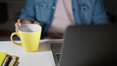 Kot ceketli genç bir adam ofis ortamındaki sarı bardaktan içiyor.