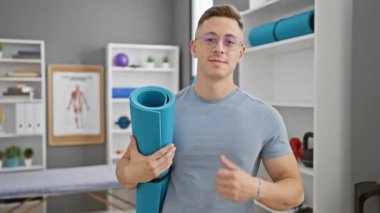Onaylayarak gülümseyen, yoga minderi tutan, rehabilitasyon kliniğinde mükemmel bir işaret gösteren pozitif İspanyol genç adam. Başarı sevincini yakalayan bu kendine güvenen adam sağlıklı ve mutlu bir yaşam tarzını somutlaştırıyor..