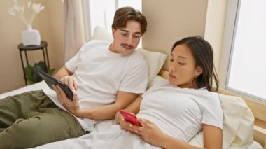 Irklar arası bir çift yatak odalarında dinleniyor, adam tablet okuyor ve kadın akıllı telefon kullanıyor..
